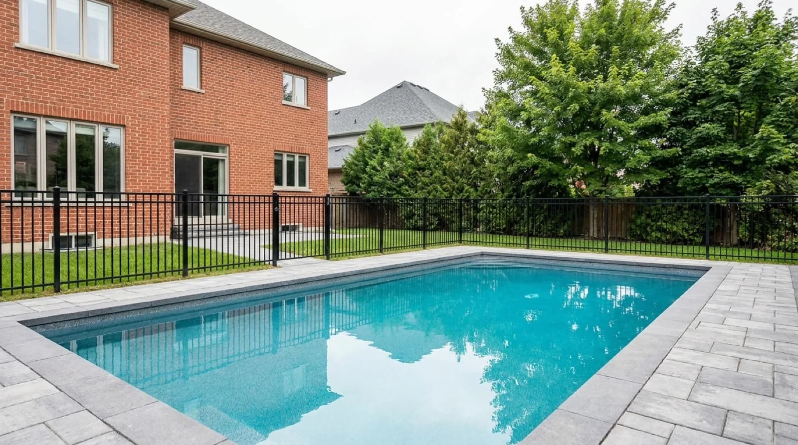 A newly finished rectangular inground pool behind a two-storey brick house in a Toronto suburb, with an interlock stone deck, a black aluminum picket fence and mature maples along the back of the lot