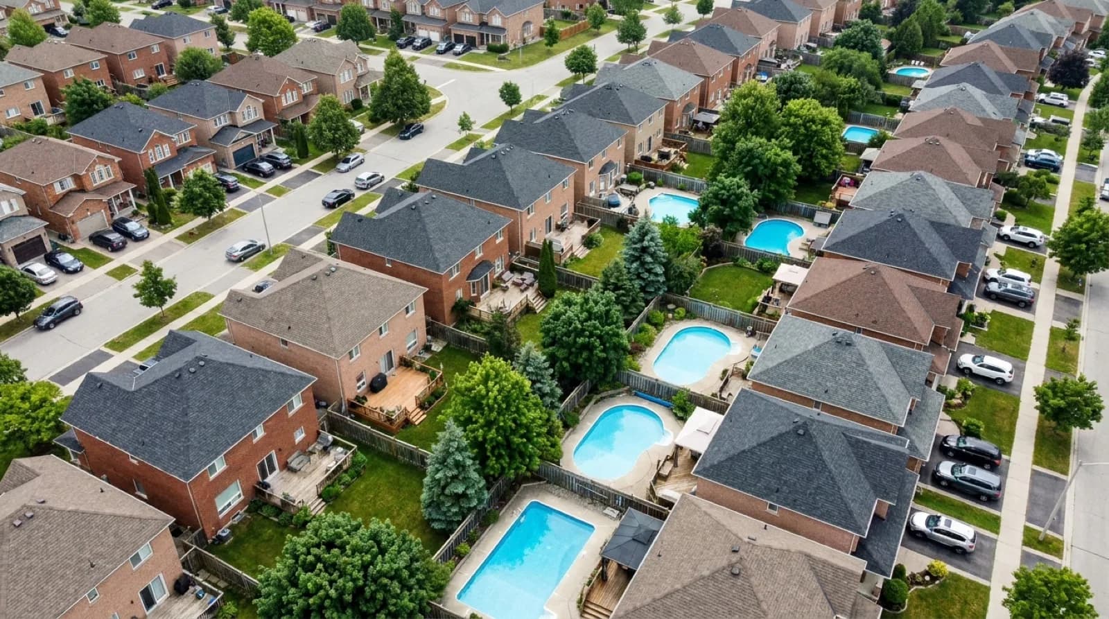 An elevated view across a suburban Greater Toronto Area neighbourhood in summer, with fenced back gardens, a handful of blue swimming pools and mature trees between the streets