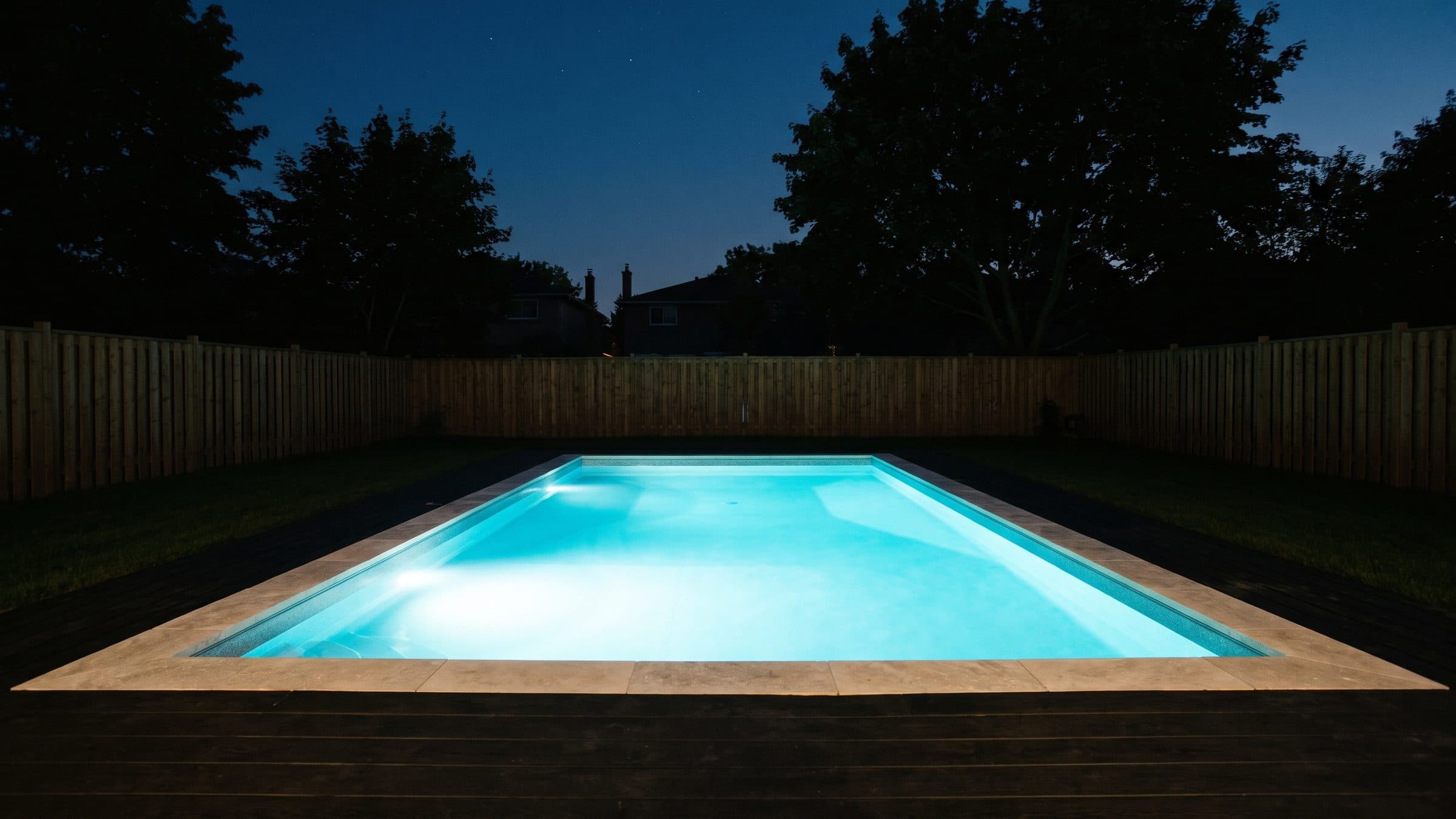 A newly finished rectangular inground pool in a fenced suburban backyard at dusk, its underwater lights on so the water is the only lit thing in the yard, with pale coping stone around the edge and the deck, lawn and cedar fence in darkness.