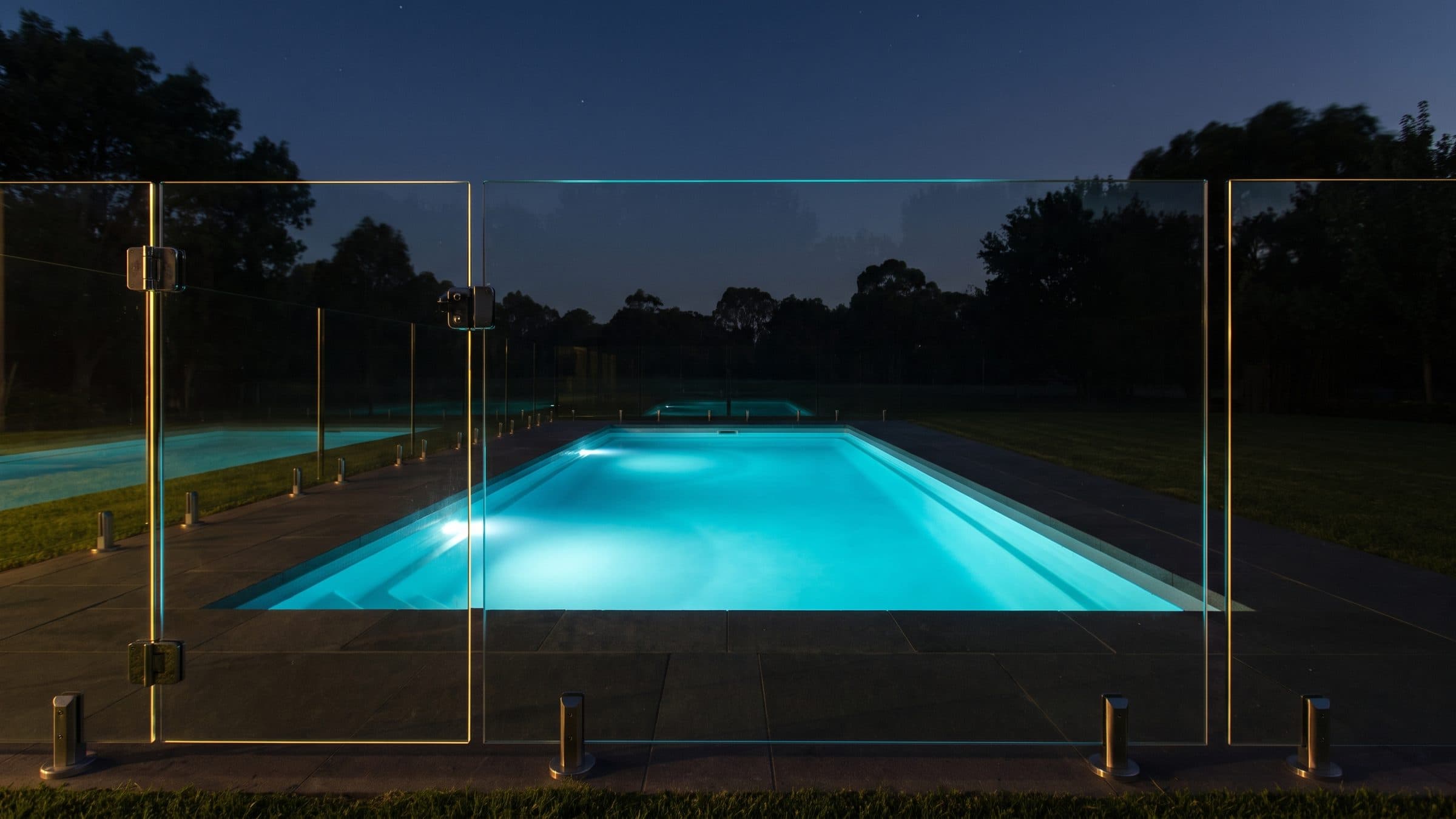 A lit pool seen at dusk from outside a frameless glass pool fence, the glass panels and their stainless spigots in the foreground, the cyan water glowing through the glass and a closed gate at the left.