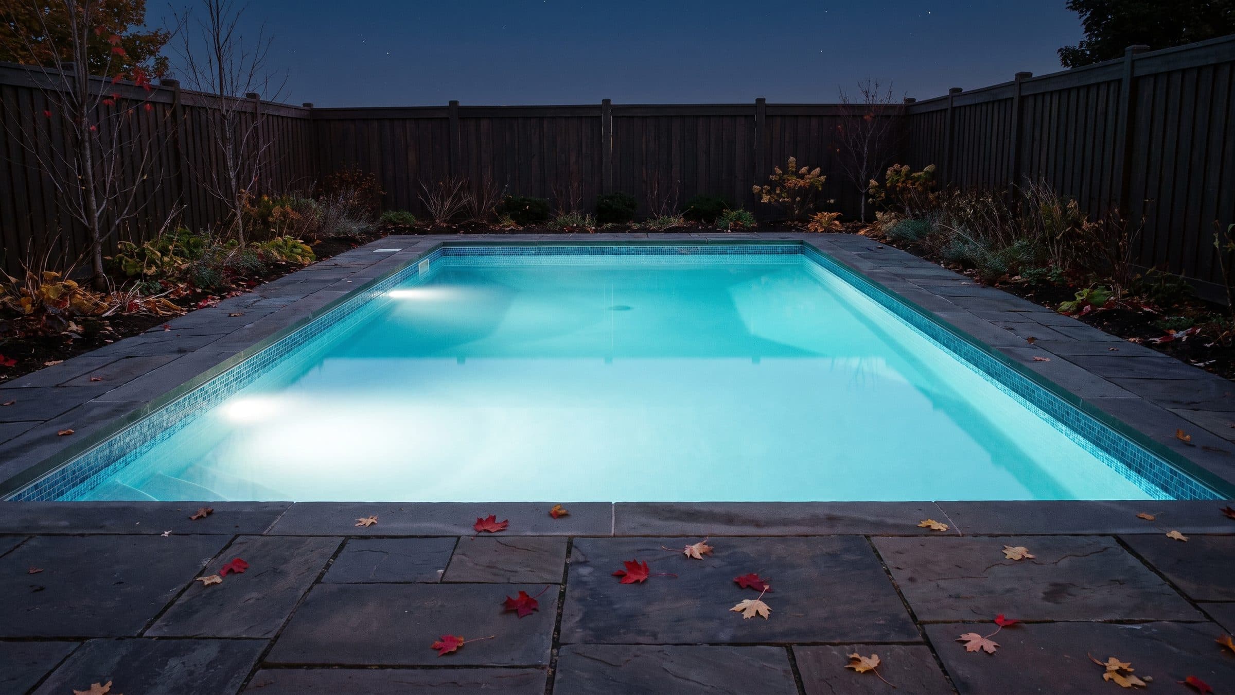 A backyard pool lit from underwater on an autumn evening, the water flat and clear enough to see the waterline tile through it, with fallen maple leaves scattered across the dark flagstone deck and none on the water.