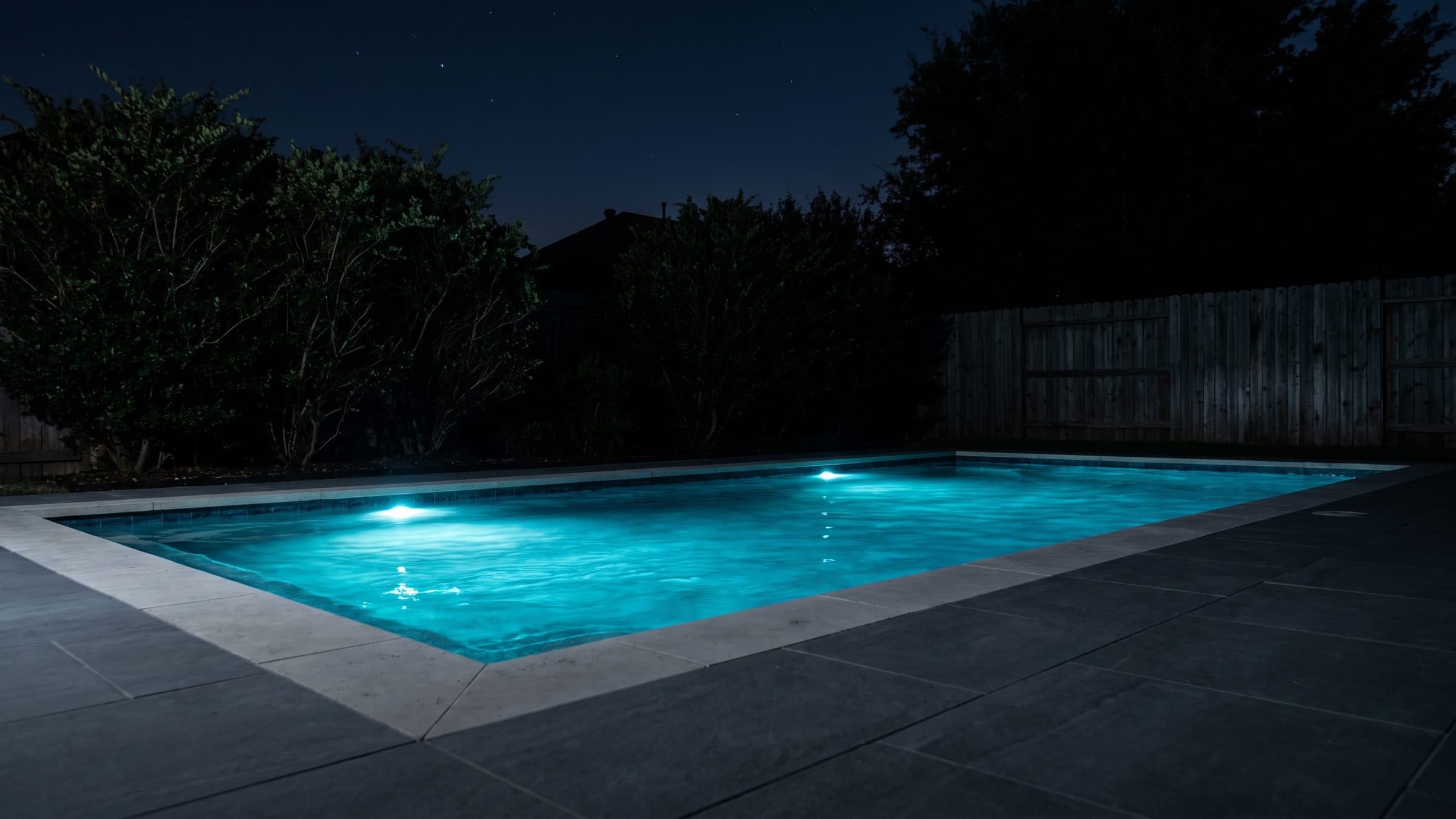 A backyard pool after dark with newly laid pale coping and a new dark stone paver deck around it, the underwater lights on, mature shrubs and a weathered board fence behind it almost black against a night sky.