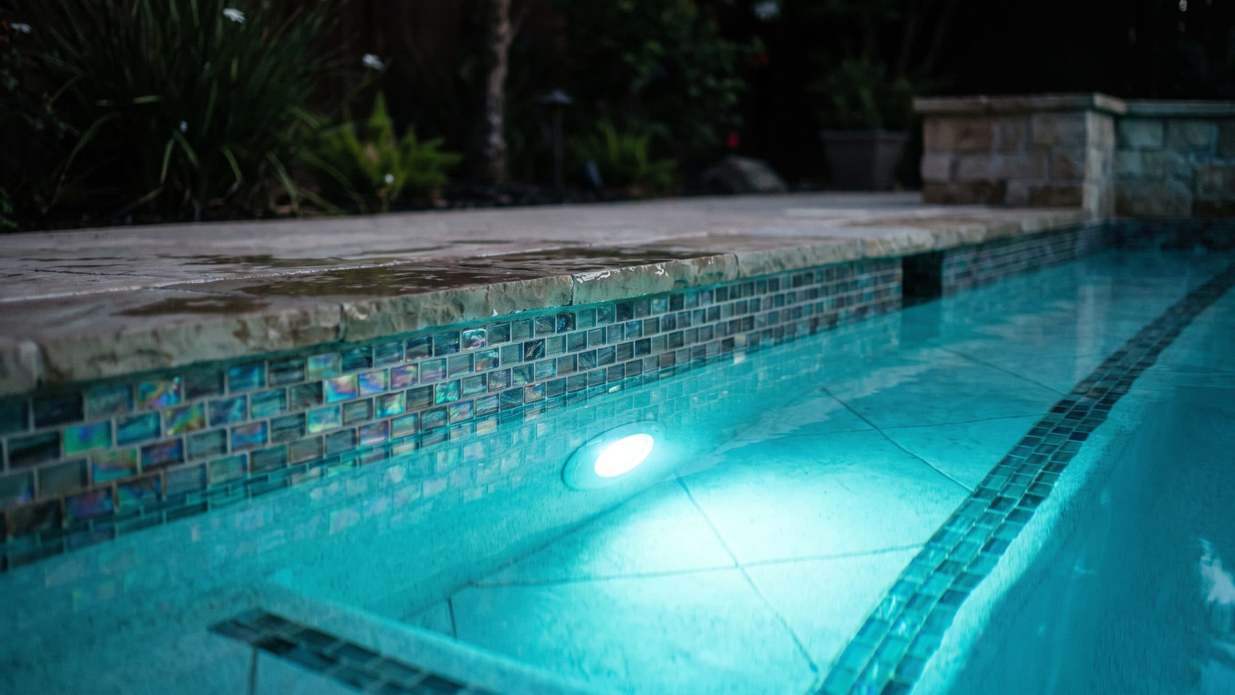 A close view along the edge of a pool at dusk showing wet limestone coping, a band of iridescent glass waterline tile, and an underwater light glowing through water clear enough to read the floor tiles beneath it.